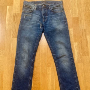 Nudie Jeans - Säljer ett par klassiska blå jeans från Nudie Jeans med snyggt slitna detaljer och kontrastsömmar på bakfickorna. Jeansen har rak passform och är i modell ”Grim Tim”.  Storleken är W30L32
