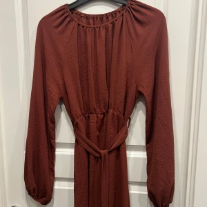 Rostbrun/röd långärmad klänning med bälte - Snygg rostbrun klänning med långa ärmar och markerad midja med knytbälte. Klänningen har en avslappnad passform och lätt rynk vid halsringningen. Perfekt för dig som gillar en enkel men trendig look.