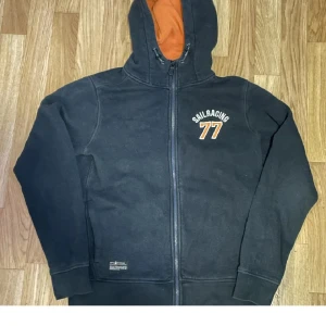 Svart Sail Racing hoodie med dragkedja - Svart hoodie från Sail Racing med orangefärgad insida på huvan och broderad text 'SAILRACING 77' på bröstet. Tröjan har hel dragkedja, snörning i huvan och långa ärmar. Perfekt för dig som gillar sportig och avslappnad stil.