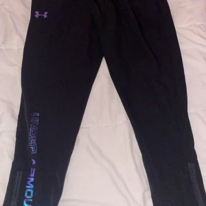 Svarta träningstights från Under Armour - Svarta träningstights från Under Armour med logga i lila på höften och text längs benet i metallicblått. Tightsen har en tight passform och är perfekta för gymmet eller löpning. Snygg sportig look med dragkedjor vid bensluten.