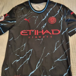 Manchester City Haaland Puma - Manchester City tröja från Puma med Haaland 9 på ryggen. Svart bas med ljusblå blixtmönster, röda detaljer och klubbmärke. Premier League-patch på ärmen.