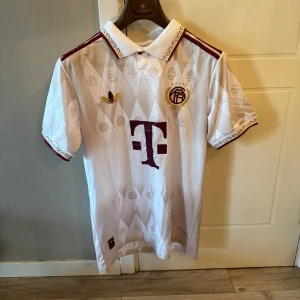 Jubeliums Bayern München Adidas fotbollströja - Snygg vit Bayern München fotbollströja från Adidas med vinröda detaljer och guldinslag. Tröjan har korta ärmar, klassisk krage med guldbroderi och diskret rutmönster. Stort T-sponsormärke på bröstet och klubbmärke med 125-årsjubileum. Tillverkad i lätt och ventilerande material.