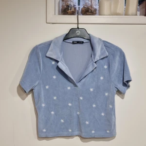 Ljusblå croppad polotopp med stjärnor - Superfin ljusblå croppad polotopp från sinsay med korta ärmar och vitbroderade stjärnor över hela toppen. Mjuk manchesterkänsla och öppen krage.