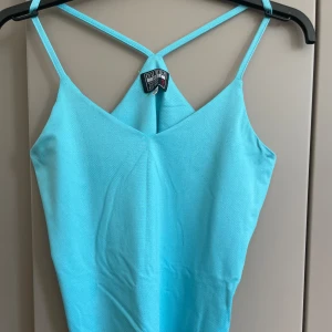 Turkos glittrig linnetopp från Guess - Supersnygg turkos linnetopp från Guess med tunna axelband som korsas i ryggen. Toppen har ett glittrigt tyg med små silverprickar och en lätt v-ringad halsringning. Perfekt för dig som vill sticka ut med en färgstark och trendig look.