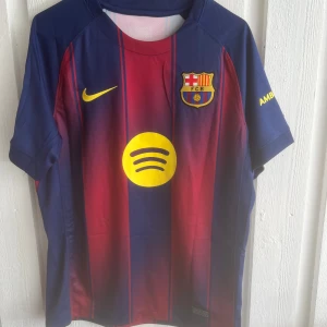 FC Barcelona Lamine Yamal matchtröja - Säljer en officiell FC Barcelona matchtröja från Nike med Lamine Yamal och nummer 10 på ryggen. Tröjan är kortärmad, har klassiska blå och röda ränder, gula detaljer och klubbmärke på bröstet. Materialet är lätt och andas, perfekt för fotboll eller supporterliv.