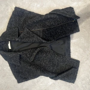 Mörkgrå omlottväst med bälte - Snygg mörkgrå väst i fiskbensmönster med omlottstängning och bälte i midjan. Västen har en markerad krage och dragkedja framtill. Perfekt lager-på-lager-plagg för kyligare dagar. Materialet känns som ullblandning och insidan är fodrad.