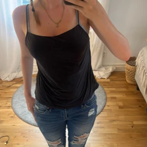 Svart linnetopp med tunna axelband - Snygg svart linnetopp med tunna axelband och avslappnad passform. Perfekt till jeans eller kjol för en enkel och stilren look. Tunt och mjukt material som känns skönt mot huden.