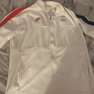 PSG x Nike vit vindjacka XL - Snygg vit PSG vindjacka från Nike i storlek XL. Jackan har dragkedja, röda och blå detaljer på ärmarna och Paris Saint-Germain-logga på bröstet. Perfekt för dig som gillar fotboll och vill ha en sportig stil. Tillverkad i polyester för en lätt och bekväm känsla. Den är i hyfsat gott skick tappade bort drack Hållaren bara☺️