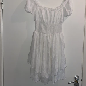 Vit offshoulder kortklänning - Superfin vit kortklänning med offshoulder och smockad midja. Klänningen har volangig kjol och puffiga korta ärmar, perfekt för sommarens alla tillfällen. Lätt och luftig i materialet, ger en romantisk vibe.