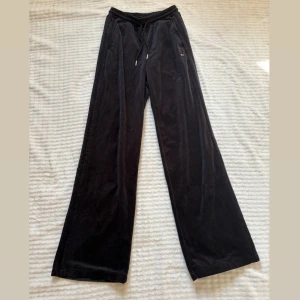 Svarta velourbyxor från Adidas - Svarta mjukisbyxor från Adidas i mjuk velour med broderad logga på höften. Byxorna har resår i midjan med snörning och raka, vida ben. Perfekta för en chill och sportig look.