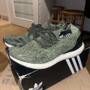 Adidas Ultraboost i grönmelerad färg - Säljer ett par Adidas Ultraboost sneakers i grönmelerad färg med stickad ovandel och vita Boost-sulor. Skorna har snörning och svart hälkappa. Perfekta för dig som gillar streetstyle och vill ha bekväma sneakers med snygg design.