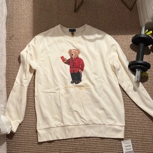 Vit Polo Bear tröja från Ralph Lauren - Vit långärmad tröja från Ralph Lauren med ikoniska Polo Bear-trycket framtill. Björnen har röd jacka och svarta byxor, vilket ger en cool och lekfull vibe. Tröjan har rund halsringning och är gjord i mjuk bomull. Perfekt för dig som gillar streetstyle och klassiska märken.