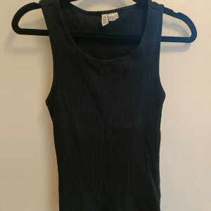 Svart ribbad linnetopp H&M - En svart ribbad linnetopp från H&M med rund halsringning och ärmlös design. Perfekt att styla med jeans eller kjol för en clean look. Mjuk och stretchig bomullskänsla som sitter snyggt på kroppen.