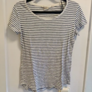 Randig vit och blå t-shirt - Snygg t-shirt med smala blå ränder på vit botten. Klassisk rund hals och korta ärmar, perfekt för en chill och enkel stil. Mjuk och lätt bomull som känns skön mot huden. Passar dig som gillar en clean och tidlös look.