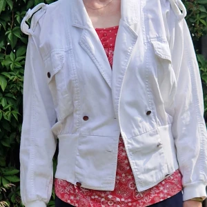 Vit jeansjacka med bred mudd nertill, fickor och bälte - Vit jeansjacka med markerad midja, stora fickor med knappar och axeldetaljer. Jackan har breda muddar nertill och på ärmsluten samt ett bälte i midjan. Lite 80-tals look.