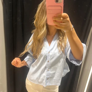 Ljusblå skjorta - Säljer en ljusblå skjorta med v-ringning och knappar framtill. Skjortan har en loose passform och vida trekvartsärmar som ger en avslappnad vibe. Perfekt att styla med jeans eller kjol för en fräsch look.