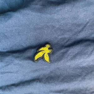 Mörkblå t-shirt från Lyle & Scott - Snygg mörkblå t-shirt från Lyle & Scott med klassisk gul fågel-logga broderad på bröstet. Tillverkad i mjuk bomull som känns skön mot huden. Perfekt för dig som gillar stilrena och tidlösa plagg med en sportig touch.