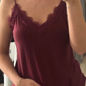 Vinröd linnetopp med spets från Amisu - Supersnygg vinröd linnetopp från Amisu med tunna axelband och v-ringning dekorerad med spets. Säljs då den ej kommer till användning.