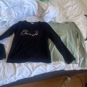 Svart och grön långärmad tröja TIKI - Två långärmade tröjor från TIKI i storlek 158/164. Ena är svart och den andra är ljusgrön, båda med snyggt guldigt tryck 'Own It' på bröstet. Tröjorna har rund hals och är gjorda i mjuk bomull, perfekta för chill dagar eller när du vill ha en enkel men cool look.