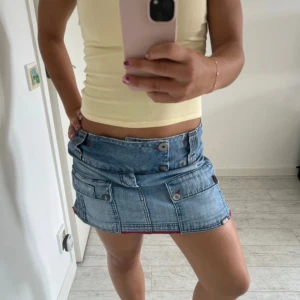 Blå Lågmidjad jeanskjol med röda detaljer - Supersnygg ljusblå jeanskjol från JGM med två stora fickor framtill och röda detaljer längs kanterna. Kjolen är kort och har bälteshällor, knappar och cool broderad logga bak. Perfekt för dig som gillar streetstyle och vill sticka ut lite extra. Midjemått 42 längd 33 cm