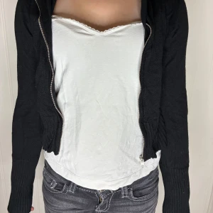 Svart stickad hoodie med dragkedja - Svart stickad hoodie med dragkedja framtill och ribbade muddar i midja och ärmslut. Modellen är croppad och har en avslappnad passform. Perfekt för lager-på-lager. Säljer på grund av att den är för liten för mig. Pris kan diskuteras❤️