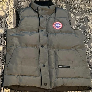 Grå dunväst från Canada Goose M - Säljer en grå dunväst från Canada Goose med hög krage och två stora fickor framtill. Västen har klassisk logga på bröstet och stängs med både dragkedja och knappar. Perfekt för lager-på-lager under kyliga dagar.