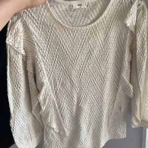 Vit blus med volang från MNG - Säljer en vit blus från MNG i storlek M. Blusen har ett snyggt stickat mönster och volangdetaljer längs axlarna. Materialet är lätt och luftigt, perfekt för vår och sommar. Rund halsringning och långa ärmar ger en chill och feminin vibe.