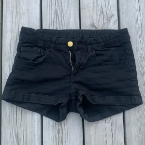 Svarta jeansshorts från Young Forever  - Svarta jeansshorts från Young Forever med klassisk femficksdesign, guldknapp och dragkedja framtill. Modellen är kort och har uppvikta benslut. Med justerbar midja. 