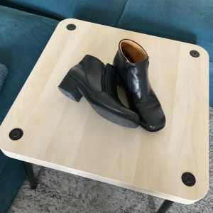Svarta stövletter i skinn med klack - Snygga svarta stövletter i skinn med låg klack och dragkedja på sidan. Klassisk design med fyrkantig tå och stilren look, perfekta till jeans eller kjol. Bekväma och lätta att ta på tack vare dragkedjan.
