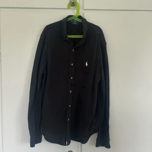 Svart skjorta från Ralph Lauren M - Skjortan har några fläckar (som syns på bilderna), dessa fläckar kan nog gå att tvätta bort. Därav säljs den så pass billigt. Svart långärmad skjorta från Ralph Lauren i lätt bomullsmaterial (featherweight mesh). Klassisk krage, knäppning framtill och vit broderad logga på bröstet.