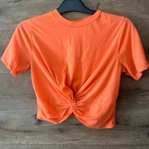 Orange croppad topp från Junkyard - Snygg orange croppad topp från Junkyard i mjuk bomull. Toppen har korta ärmar och en cool twistad knut framtill som ger en trendig look. Perfekt till jeans eller kjol för en färgstark outfit.