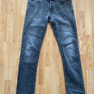 Grå jeans från Nudie Jeans  - Säljer ett par grå jeans från Nudie Jeans Co med klassisk femficksdesign och svarta broderade detaljer på bakfickorna. Jeansen har en rak passform och är tillverkade i mjukt denimtyg. Perfekta för dig som gillar en avslappnad men stilren look.