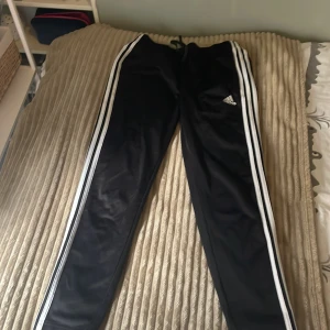 Svarta Adidas mjukisbyxor med ränder - Svarta mjukisbyxor från Adidas med klassiska vita ränder längs sidorna och Adidas-logga på låret. Byxorna har dragsko i midjan och är gjorda i ett glansigt syntetmaterial. Perfekta för chill eller träning.