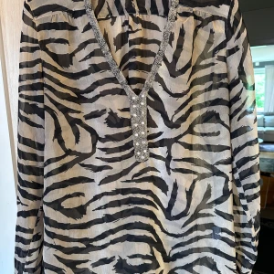 Zebramönstrad blus med paljetter strl: M - Snygg transparent blus från RoseBud i beige och svart zebramönster. V-ringad med glittriga paljettdetaljer längs halsringningen och knäppning framtill. Långärmad och lätt, perfekt för dig som vill sticka ut med coola detaljer och djurmönster.