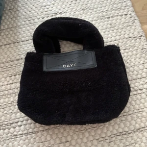 Svart teddy handväska från DAY ET - Supermjuk svart handväska från DAY ET i teddy-material med korta handtag och snygga detaljer i svart skinnimitation. Väskan har magnetlås och innerfack med dragkedja, perfekt för att hålla ordning på dina grejer. Aldrig använd men prislapp borttagen. Pris kan diskuteras.
