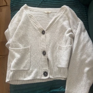 Beige kofta från H&M med stora knappar - Supermysig beige kofta från H&M med stora svarta knappar och två fickor framtill. Koftan har v-ringning, ribbade muddar och är stickad i ett mjukt material. Perfekt att slänga på sig när det är lite kyligt ute.