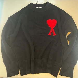Svart stickad tröja från Ami Paris - Svart stickad tröja från Ami Paris med röd hjärtlogga och bokstaven A på bröstet. Tröjan har ribbade muddar, rund hals och en loose fit. Perfekt för dig som gillar stilrena plagg med en twist. Materialet är mjukt och känns lyxigt.Priser kan diskuteras vid snabb affär.