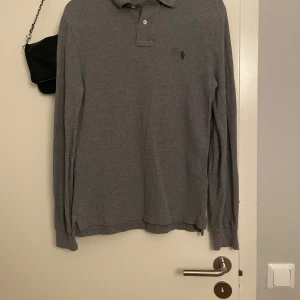 Grå långärmad pikétröja från Polo Ralph Lauren - Klassisk grå långärmad pikétröja från Polo Ralph Lauren med liten broderad logga på bröstet. Tröjan har krage, knappar vid halsen och är gjord i mjuk bomull. Perfekt för en clean och stilren look.