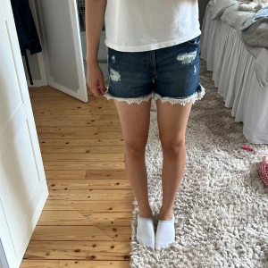 Blå jeansshorts med spetskant - Snygga blå jeansshorts med slitna detaljer och vit spetskant längst ner. Modellen är midwaist och har klassisk femficksdesign. Perfekta för sommarens alla äventyr och ger en cool och avslappnad vibe.