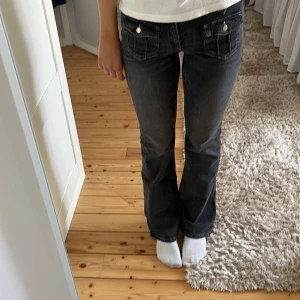 Svarta bootcut jeans med fickdetaljer - Snygga svarta jeans med bootcut passform och coola fickor framtill med lock och knappar. Jeansen har låg midja och är tillverkade i klassiskt denimtyg. Perfekta för dig som gillar en avslappnad men ändå trendig look. Är inte använda💓 Hör av dig vid frågor!