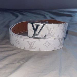 Vit monogram skärp - Snyggt vitt bälte i skinn med klassiskt monogrammönster och stor silverfärgad spänne. Bältet har en stilren look . Perfekt accessoar för att lyfta din outfit.