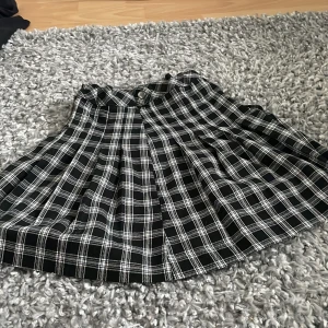 Svartvit rutig kortkjol med knappar - Supersnygg svartvit rutig kortkjol med plisserad design och knappar framtill i midjan. Kjolen har en klassisk skolstil och är perfekt för dig som gillar preppy vibes. Materialet känns mjukt och följsamt, och mönstret är tidlöst. Passar dig som vill sticka ut med en cool look.