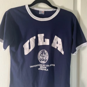 Blå ULA t-shirt/top  - Snygg mörkblå t-shirt/top med vit text och emblem från Universidad de Los Andes, Venezuela. T-shirten har vita ribbade kanter vid hals och ärmslut, klassisk rund hals och korta ärmar. Perfekt för dig som gillar retro college-vibe. Kort modell.  Som en stl M i tight modell (stl 14)