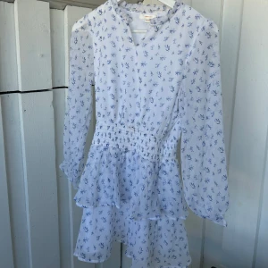 Blommig vit kortklänning Gina Tricot - Supersöt vit kortklänning från Gina Tricot med blått blommigt mönster. Klänningen har långärmade transparenta ärmar, v-ringad hals och smock i midjan för en snygg passform. Volangdetaljer på kjolen ger extra flow. Perfekt för sommarens alla tillfällen! Lite miss färgningar som de syns på bilderna, men går lätt bort med ex vanish🥰
