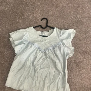 Ljusblå blus med spetsdetalj - Söt ljusblå blus med korta volangärmar och rund hals. Framtill finns en dekorativ spetsdetalj i samma färg som ger en extra touch. Perfekt för dig som gillar romantisk och feminin stil.