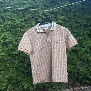Beige mönstrad Lacoste t-shirt - Snygg beige t-shirt från Lacoste med brunt geometriskt mönster och klassisk krage med ljusblå och bruna detaljer. Ikonisk krokodillogga på bröstet och bruna knappar. Skön bomullskvalitet och korta ärmar, perfekt för chill dagar.
