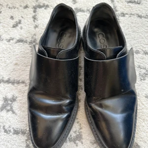 Svarta skinnloafers från COS - Stilrena svarta loafers från COS i blankt skinn med bred rem över vristen och rund tå. Snygg minimalistisk design och robust sula. Perfekta för dig som gillar en clean och modern look.