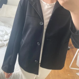 Svart kappa med huva och knappar - Stilren svart kappa med huva och tre knappar framtill. Jackan har raka linjer, två snedställda fickor och är helsvart. 