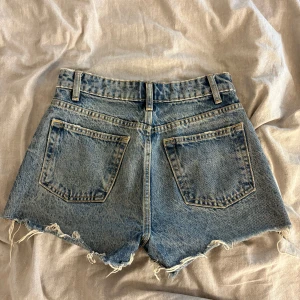 Blåa jeansshorts från Zara - Snygga blå jeansshorts från Zara med medelhög midja och råa, fransiga benslut. Klassisk femficksmodell med bälteshällor och knappgylf. Perfekta för varma dagar och ger en avslappnad vibe.