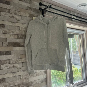 Grå hoodie från H&M Divided - Säljer en ljusgrå hoodie från H&M Divided i storlek S. Tröjan har huva med snörning, dragkedja framtill och två stora fickor. Perfekt för chill dagar och enkel att matcha med jeans eller mjukis. Mjuk bomullskänsla och långärmad modell.